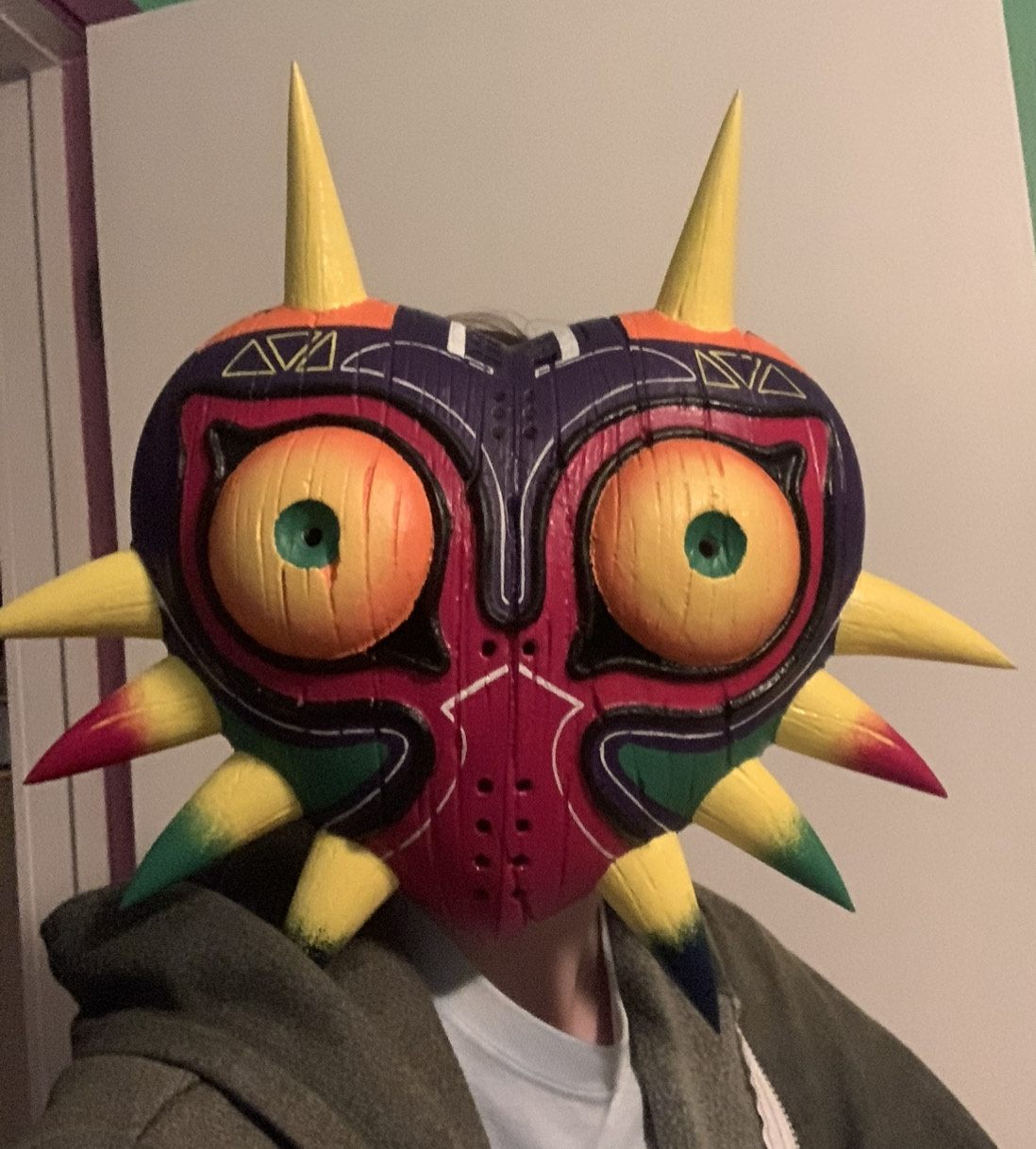 A photo of a person wearing a replics of majora's mask, a colorful heart shaped mask with large yellow eyes, no mouth or nose, and spikes on the side and two horns