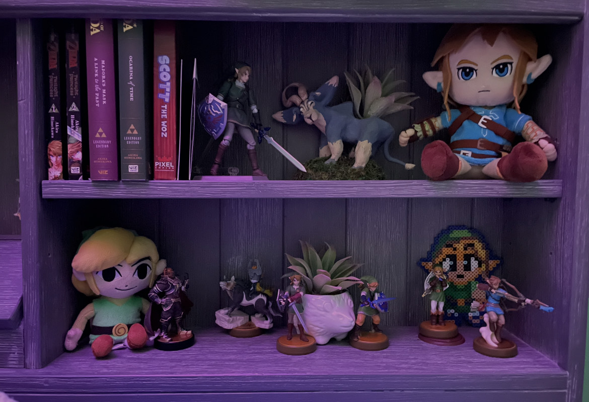 A shelf containg various different Zelda merch including 2 plushes, 4 mangas, 7 figures, and a planter